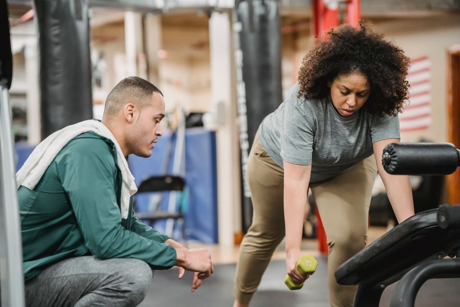 Multiracial coach and woman doing exercises with dumbbell and gym equipment on blurred background of sport club